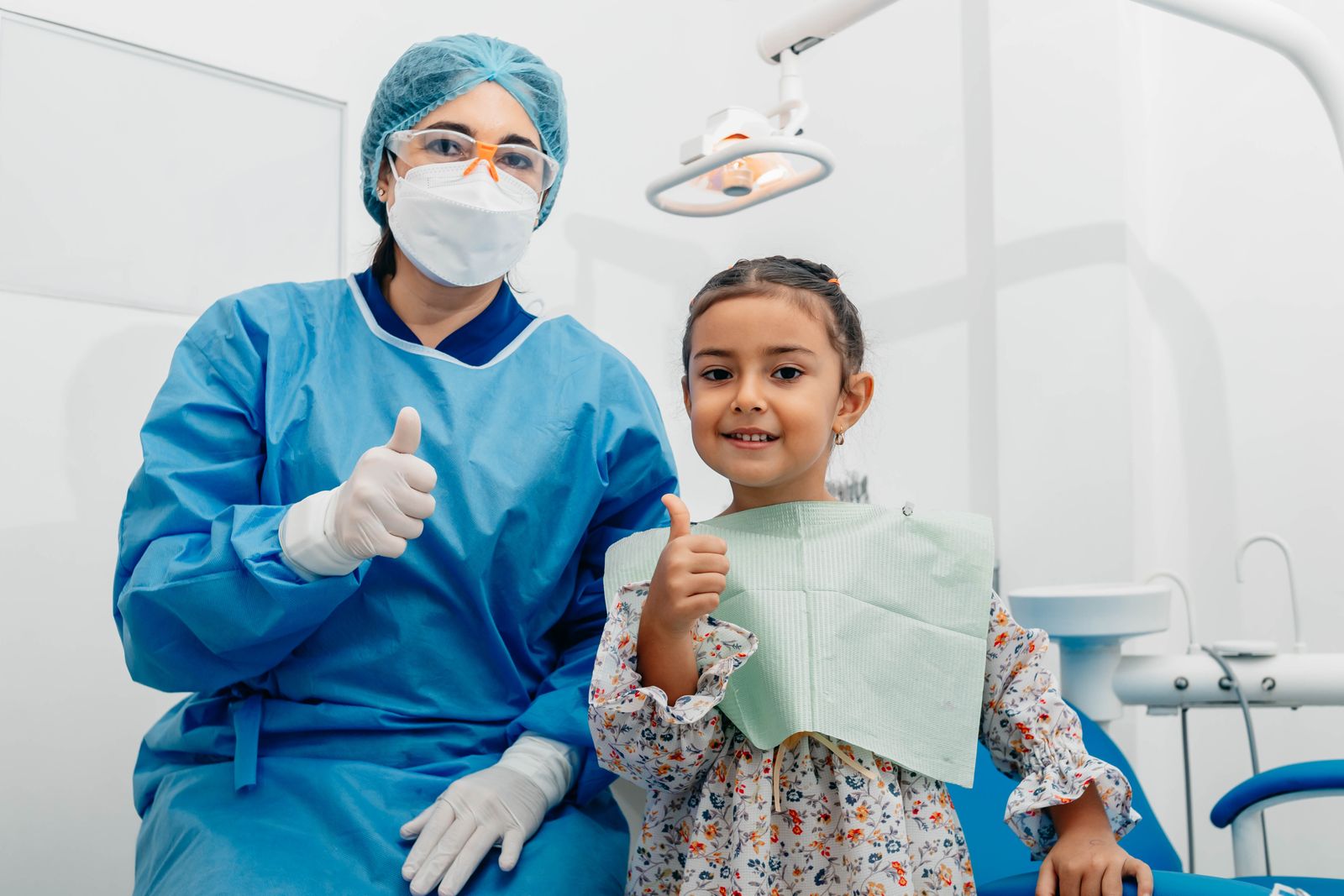 Lächelnde Zahnärztin mit kleiner Patientin in der Praxis, beide zeigen den Daumen nach oben